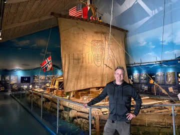 The Kon-Tiki is a balsa wood raft with a rectangular shape with a large square sail displaying a stylized face design. The raft is made of giant balsa logs tied together with rope on top of which sits a small cabin. The ship is decorated with various flags, including Norway, Peru and the United States.